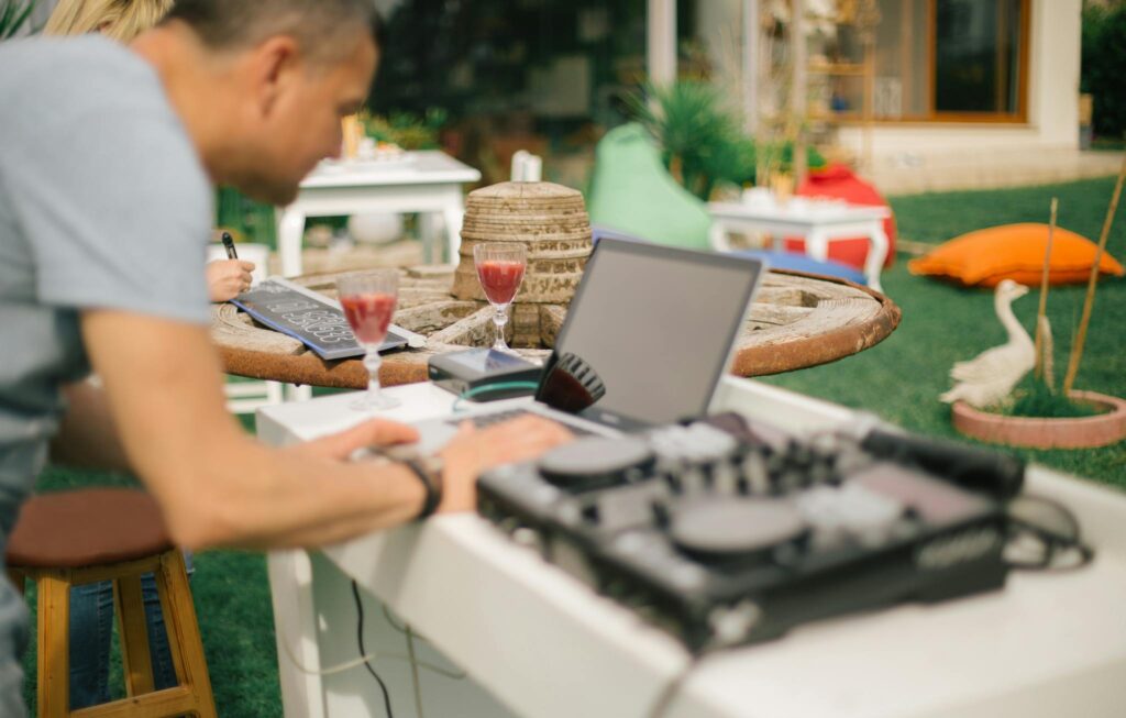 DJ using laptop and mixer outdoors on a sunny day, with drinks on the table.